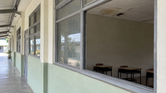 Vista desde un pasillo hacia un aula con una ventana sin vidrio, pupitres y manchas de humedad en el cielo raso.