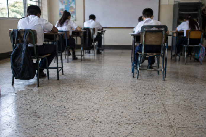 Estudiantes de espaldas trabajando en sus pupitres, con el piso del aula en primer plano.